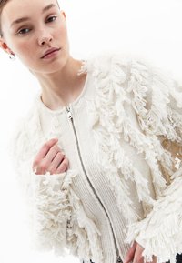 Cardigan a maglia color avorio con dettagli in frange testurizzate, chiusura con zip davanti e polsini a coste. Tessuto morbido e peloso con un aspetto a strati.