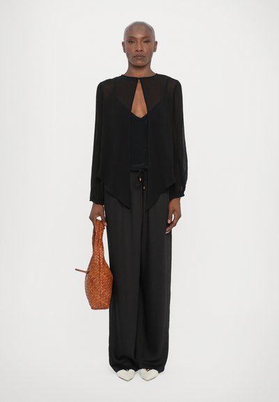 Black sheer blouse with a cutout neck, paired with loose black pants. Accessories include a woven brown bag and cream-colored shoes.