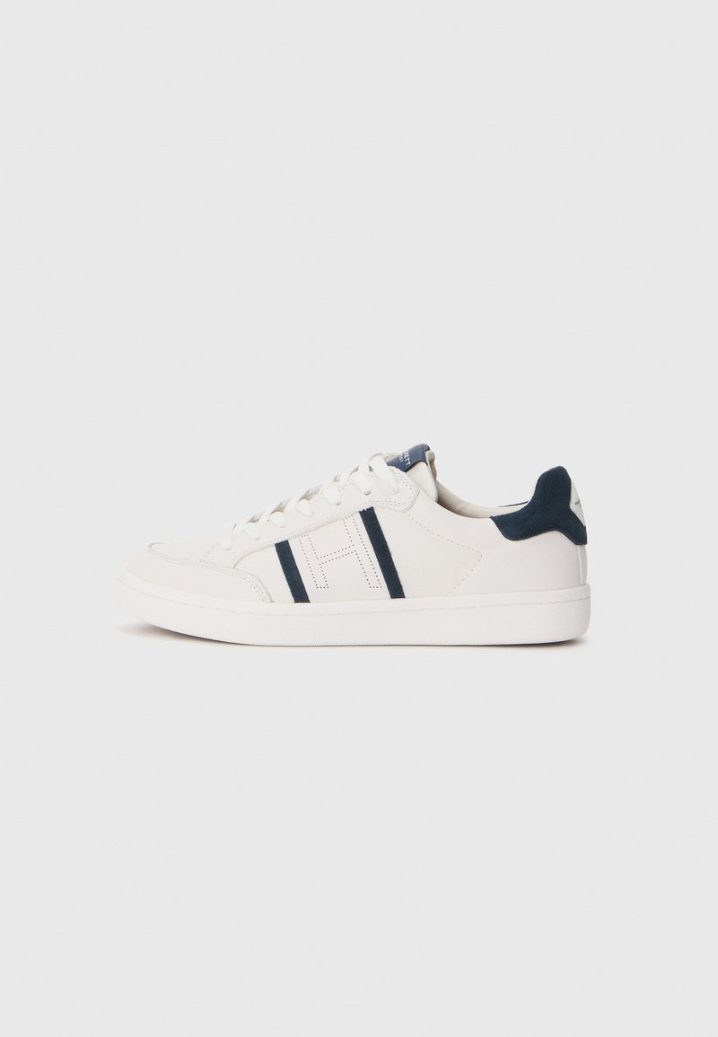 Baskets basses blanches avec un talon en daim navy et des rayures navy sur le côté, lacets blancs et semelle blanche plate sur fond uni.
