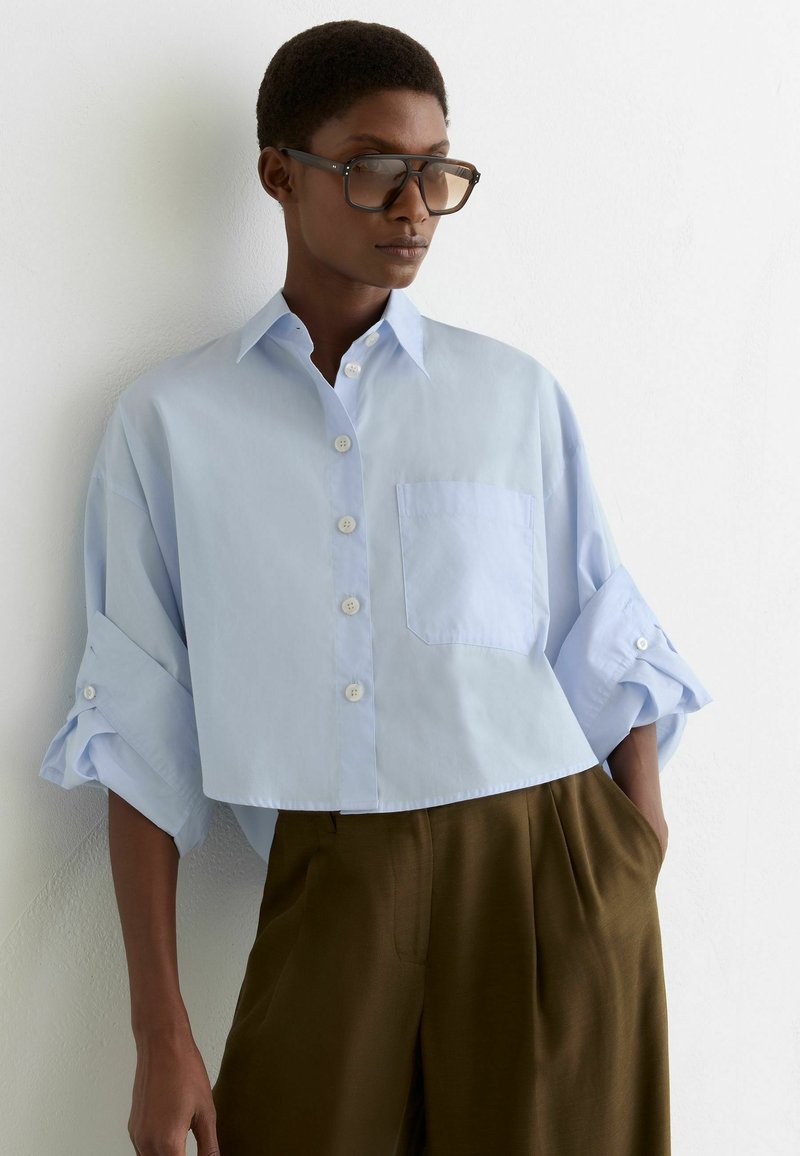 Rövid hajú személy, nagy szemüveget visel, túlméretezett világoskék gombos ingben, felhajtott ujjakkal és barna, széles szárú nadrágban, a falnak dőlve.