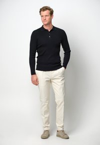 Dunkelblaue gestrickte Poloshirts mit kurzem Kragen, kombiniert mit cremefarbenen Chinos und hellbeigen Sneakers, vor einem schlichten Hintergrund stehend.