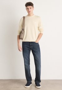 Beige sweater with long sleeves, dark blue jeans, and navy sneakers. The man carries a small beige shoulder bag. Plain background.