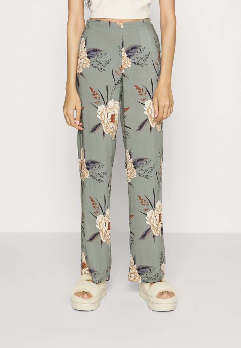 Femme portant un pantalon large taille haute vert à motif floral et des sandales crème fourrées, debout devant un fond blanc uni.