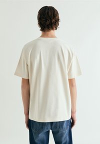 Persona con cabello castaño de longitud media, vistiendo una camiseta lisa de manga corta, holgada y color blanco roto, y pantalones vaqueros azules, vista desde atrás.