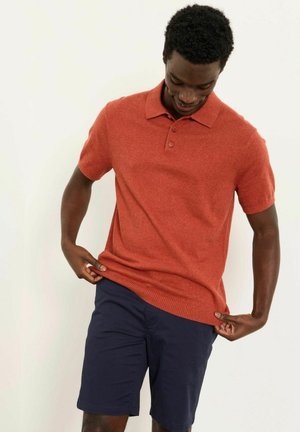 Vyras dėvintis rūdžių spalvos trumpomis rankovėmis polo marškinėlį ir tamsiai mėlynas šortus, reguliuojantis marškinėlių apačią ir žvelgiantis žemyn.