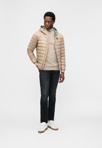 Άνδρας με μπεζ μπουφάν puffer πάνω από μπεζ πουλόβερ, σκούρο τζιν και λευκά αθλητικά παπούτσια, στέκεται με το ένα χέρι στην τσέπη μπροστά σε λευκό φόντο.