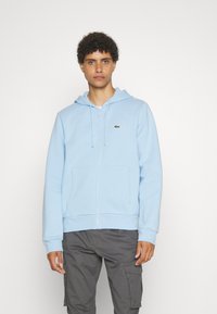 Šviesiai mėlynas zip-up džemperis iš minkštos medžiagos, su priekinėmis kišenėmis, kapisonu su raišteliais ir mažu žaliu logotipu ant krūtinės. Derinamas su pilkais cargo kelniais.