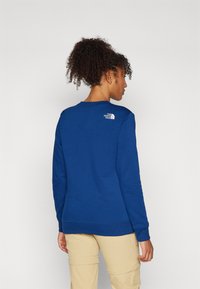 Kék pulóver puha anyagból, kerek nyakkal és bordázott mandzsettákkal. A The North Face logó fehér hímzéssel található a hátsó oldalon.