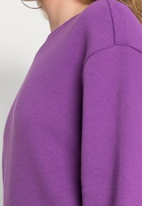 Sudadera morada con cuello redondo, material de algodón suave y textura lisa. Presenta un diseño ajustado y costuras prominentes en los hombros.