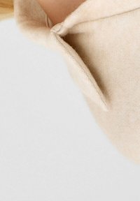 Mascherina in tessuto beige morbido con design strutturato e bordo curvo. Presenta un'anello auricolare su un lato per un'indossabilità sicura.