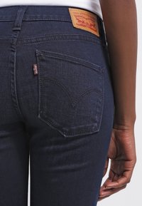 Jeans de mezclilla oscura con un bolsillo trasero cosido que presenta un diseño curvado distintivo. Una etiqueta de cuero marrón en la cinturilla muestra los detalles de la marca.