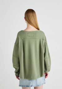 Kvinne med langt blondt hår iført en løs, strukturert olivenfarget genser og lys blå denimskjørt, stående vendt bort fra kameraet mot en ensfarget bakgrunn.