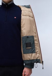 Giacca imbottita beige con un maglione di lana blu navy sotto, con una tasca verde a contrasto e dettagli in zip. Polsini a coste texturizzati.