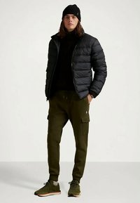 Casaco puffer preto por cima de um suéter preto, calças cargo oliva com bolsos e sapatilhas de desporto verdes. Acessório: gorro preto.