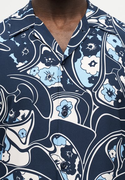 Άτομο με σκουρόχρωμο δέρμα που φοράει πουκάμισο με κουμπιά σε σκούρο μπλε και ανοιχτό μπλε με λουλουδάτο μοτίβο και γιακά που ανοίγει πλατιά.