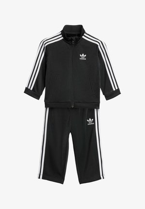 Černý Adidas teplákový komplet s bílými pruhy. Spodní zapínací bunda má stojáček; kalhoty mají elastický pas a boční pruhy.