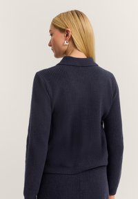 Blonde Frau trägt einen dunkelblauen, gerippten, langärmligen Pullover und einen passenden Rock, mit dem Rücken zugewandt, mit sichtbaren silbernen Ohrringen.