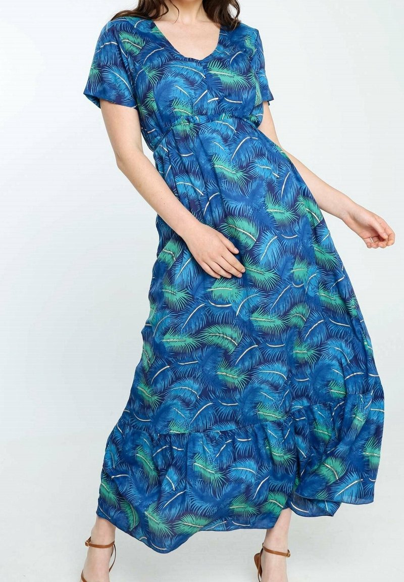 Femme portant une robe longue bleue avec un motif de feuilles de palmier vertes et blanches, manches courtes, et des sandales marron à bride cheville, posant sur fond blanc.