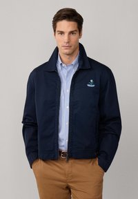 Giacca a zip blu navy con colletto rialzato, tasche laterali e logo ricamato sul petto, indossata sopra una camicia a strisce blu chiaro.