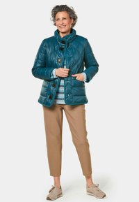 Türkis gesteppte Jacke mit hohem Kragen, ausgestattet mit schwarzen Knöpfen und diagonaler Naht. Getragen über einem hellblauen Pullover und beigen Hosen.