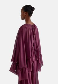 Top in chiffon color marsala con volant sovrapposti, dotato di scollatura rotonda e dettaglio con bottone sul retro. Design leggero e fluido.