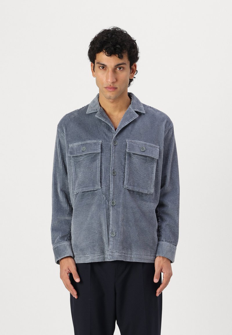 Camisa de pana azul con dos bolsillos frontales, diseño de botones, cuello extendido y mangas largas. Presenta una textura acanalada en toda su superficie.