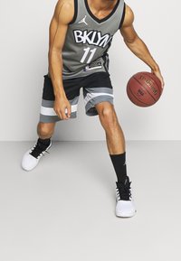 Maglia da basket grigia con "BKLYN" e numero "11", abbinata a pantaloni neri con strisce grigie e bianche. Il giocatore dribbla un pallone da basket.