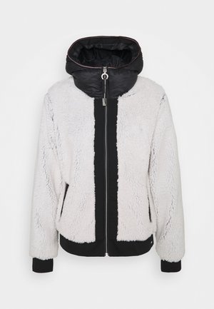 Falsche Pelzjacke in Hellgrau mit einem schwarzen Panel, ausgestattet mit einem Frontreißverschluss, elastischen Bündchen und einer Kapuze. Strukturierte Außenseite und glattes Futter.