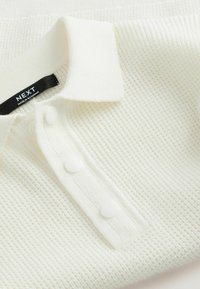 Polo shirt bianco lavorato a maglia con un motivo testurizzato, patta con tre bottoni e colletto piatto. Etichetta con la scritta "NEXT, MADE IN MYANMAR."