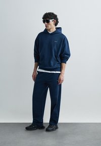 Sudadera con capucha azul marino y pantalones deportivos a juego hechos de tela suave, con un ajuste relajado y una marca sutil. Llevados con gafas de sol oscuras.