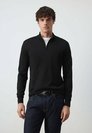 Hombre con suéter negro con cremallera hasta el pecho sobre camisa blanca y vaqueros azul oscuro, de pie contra un fondo liso y claro.