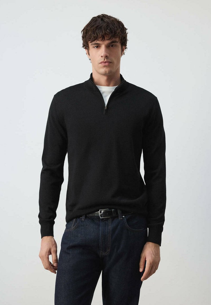 Homme portant un pull noir à fermeture éclair quart de zip sur une chemise blanche et un jean bleu foncé, debout devant un arrière-plan clair uni.