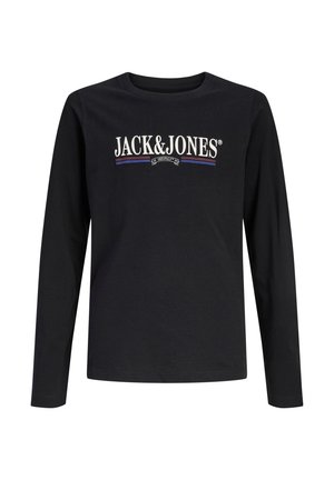 Chłopiec stoi, ubrany w czarną koszulkę z długim rękawem Jack & Jones, szerokie niebieskie dżinsy i białe trampki na jednolitym tle.