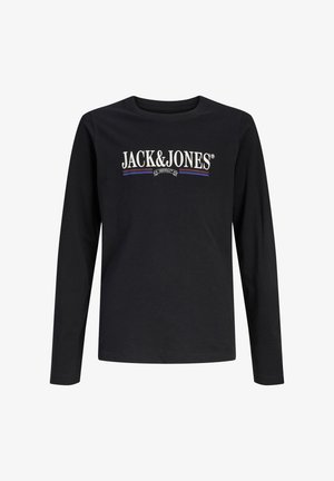 Chłopiec stoi, ubrany w czarną koszulkę z długim rękawem Jack & Jones, szerokie niebieskie dżinsy i białe trampki na jednolitym tle.
