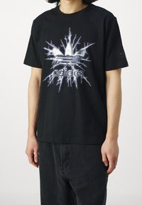 Black cotton t-shirt featuring a large silver Adidas logo with lightning graphic. Short sleeves, relaxed fit, simple round neckline.