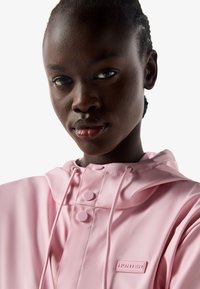 Nahaufnahme einer Person, die einen pinkfarbenen Hunter-Regenmantel mit Kapuze trägt, mit sichtbaren Knöpfen und Kordelzügen, vor einem einfarbigen weißen Hintergrund.