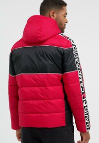 Rote und schwarze Daunenjacke mit Kapuze. Merkmale sind horizontale Farbblöcke und weiße Akzentlinien. Textlogo entlang des Ärmels. Strukturierter Stoff.