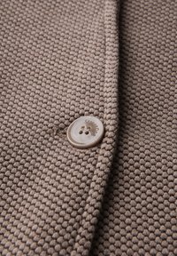 Tissu texturé marron avec un motif en nid d'abeille, comportant un bouton beige avec un logo. Le gros plan montre les détails du tissu et les coutures.
