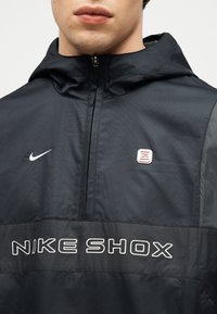 Κοντινό πλάνο ενός ατόμου που φοράει μαύρο μπουφάν Nike Shox με κουκούλα, μισό φερμουάρ και λευκό λογότυπο στο στήθος.