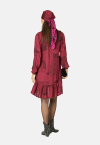 Weinrotes Kleid mit schwarzem Punktmuster, gerafftem Saum, langen Ärmeln, kombiniert mit einem rosa Schal und einer schwarzen Clutch. Lange schwarze Strumpfhosen und Ankle Boots.