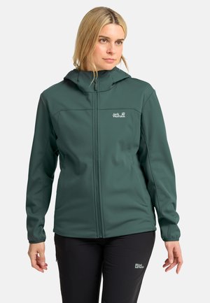 Vrouw draagt een donkergroene capuchonvest met ritssluiting en zwarte broek met outdoor merklogo's op beide kledingstukken, staand tegen een effen achtergrond.