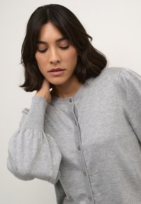 Cardigan gris clair en tricot doux, avec un col rond, une fermeture à boutons à l'avant et des manches longues bouffantes avec des poignets côtelés.