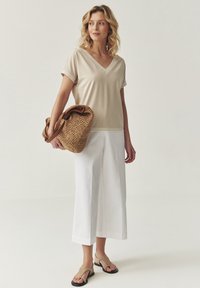 Top beige de cuello en V con mangas cortas, pantalones blancos de pierna ancha y un bolso de paja trenzada con asas de cuero marrón. Sandalias planas completan el atuendo.