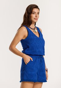 Plava bezrukavna majica s V-izrezom i teksturiranim uzorkom, uparen s odgovarajućim šortsevima koji imaju elastičnu traku u struku i resastim rubom.