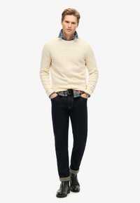 Crème textuur trui over een geruit shirt, donkere denimbroek en zwarte leren laarzen met opgerolde manchetten. Schone achtergrond.