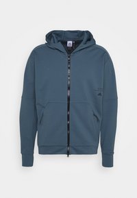 Felpa con cappuccio e zip in verde scuro, realizzata in tessuto testurizzato. Presenta due tasche frontali e una zip nera, con accenti del logo.