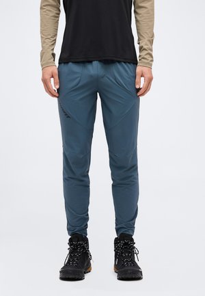 Homme portant un pantalon de sport bleu avec des bottes de randonnée noires, debout, les mains détendues le long du corps sur un fond uni.