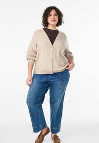 Bēšas adītas kardigāns ar V veida izgriezumu, sarullētām piedurknēm un trim pogām, apvienots ar ziliem taisna griezuma džinsiem un brūnām kurpēm.