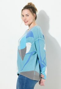 Stickad tröja i ljusblå med abstrakta geometriska mönster i mörkare blått, grått och vitt. Ribbad nederkant och avslappnad passform.