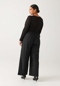 Femme en haut à manches longues en dentelle noire, pantalon noir à jambes larges et bottes à talons noires, se tenant de côté contre un fond blanc uni.